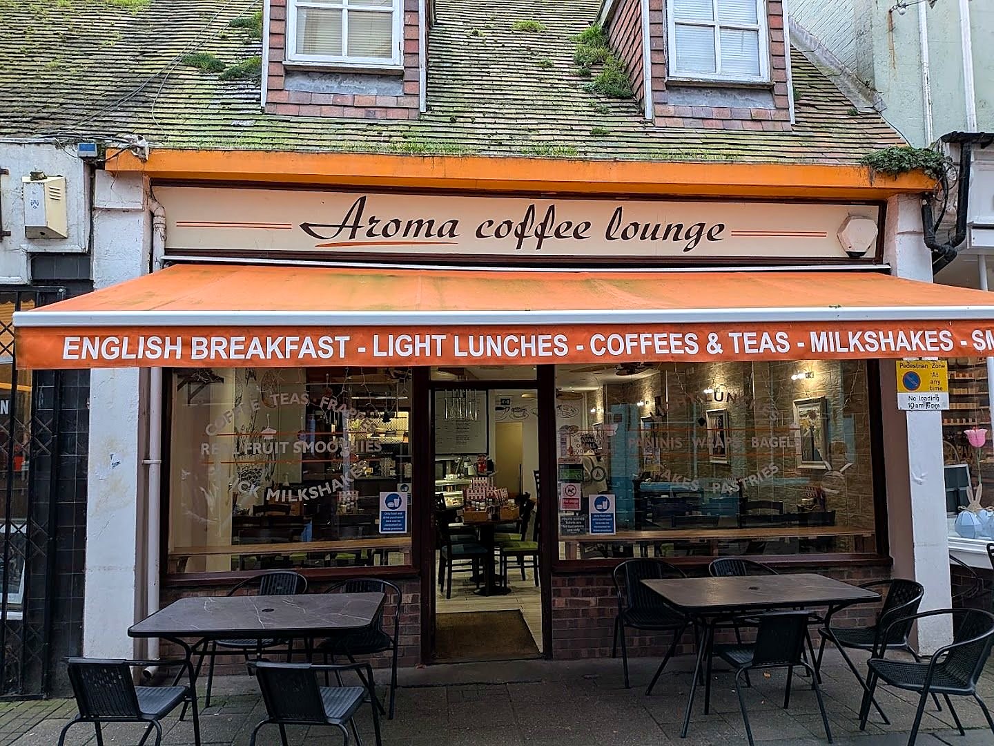Aroma Coffee Lounge: a cream sign in script lettering above an orange awning reading English Breakfast, Light Lunches, Coffees & Teas, Milkshakes, with black tables and chairs on the pavement